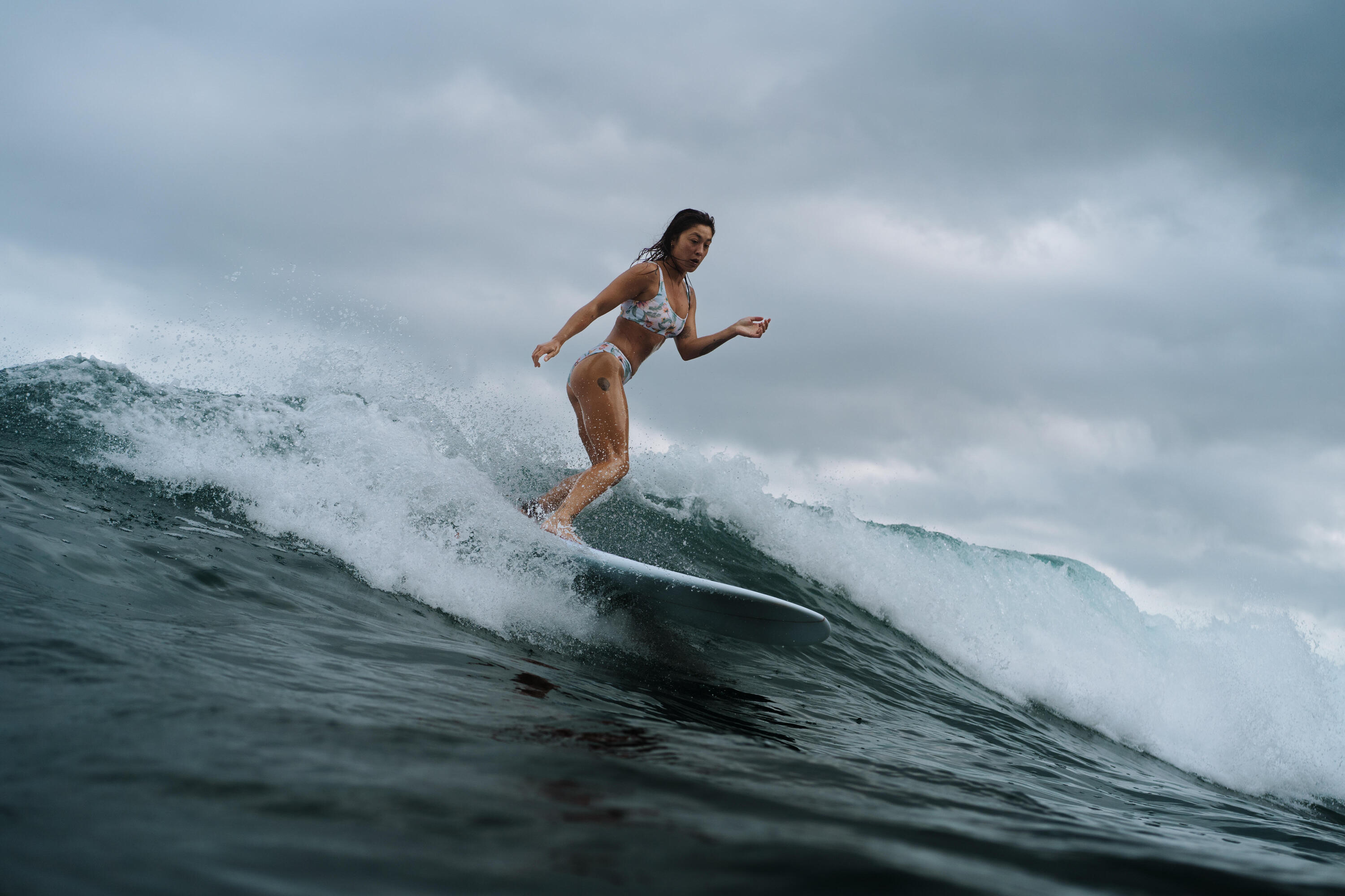 Eine Surferin in einem hellen Bikini reitet eine große, brechende Welle. Die Aufnahme zeigt sie in einer dynamischen Bewegung auf dem Board unter einem bewölkten, dramatischen Himmel.