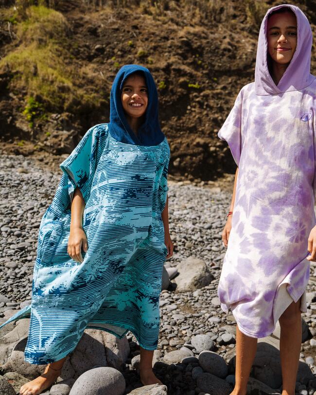 Zwei Personen in farbigen Ponchos, blau und lila, stehen auf einem steinigen Strand vor einer felsigen Klippenkulisse.