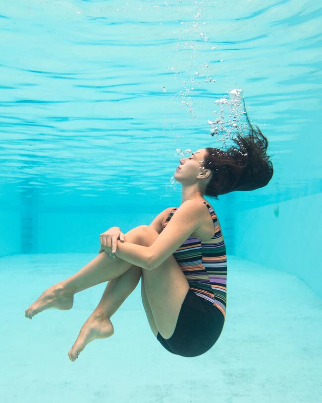 Frau taucht im Schwimmbecken unterwasser, zieht Beine an, Badeanzug gestreift