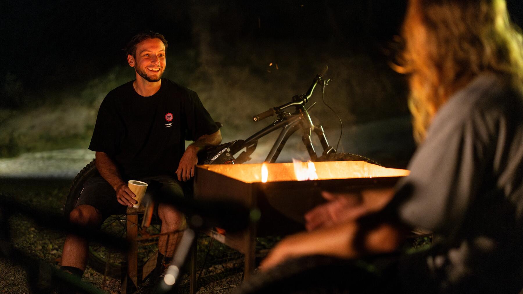 Personen sitzen bei Dunkelheit auf Campingstühlen vor einem Lagerfeuer