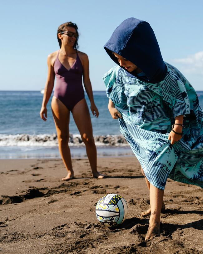 Kind im blauen Poncho spielt mit buntem Ball am Sandstrand, Frau im Badeanzug lacht, blauer Himmel, Meer im Hintergrund.