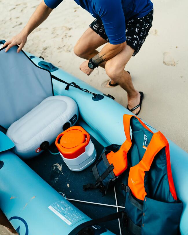 Mann im blauen Shirt mit Sonnenbrille neben blauem Schlauchboot und orange-blauen Schwimmwesten am Sandstrand.