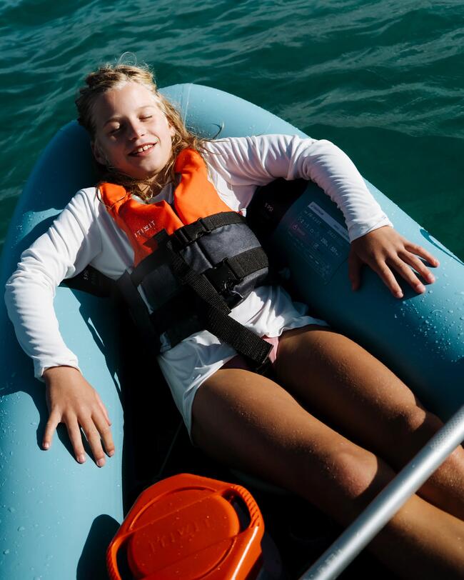 Kind in blauem Schlauchboot, trägt orangefarbene Schwimmweste, entspannt auf türkisfarbenem Wasser, Sonnenschein.