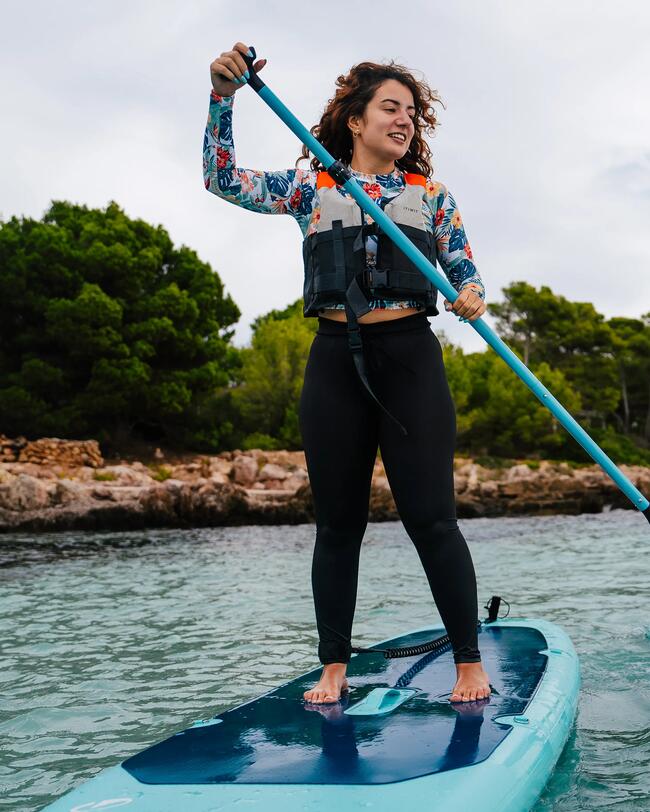 Frau paddelt auf türkisfarbenem Paddleboard, trägt bunte Kleidung und Schwimmweste, umgeben von klarem Wasser und grüner Küste.