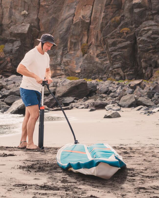 Mann pumpt blaues Paddleboard am felsigen Strand auf, trägt weißen Hut und T-Shirt. Schwarze Felsen im Hintergrund.