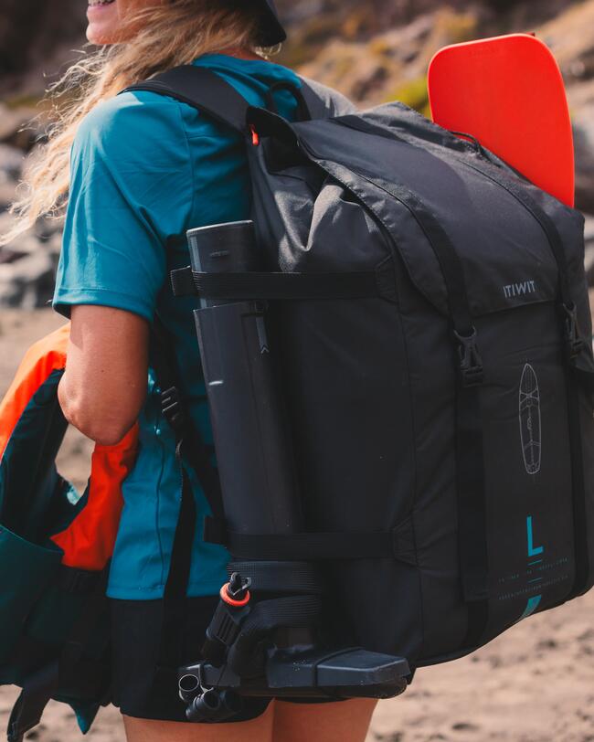 Frau mit blauem Shirt trägt großen schwarzen Rucksack, orangene Paddel sichtbar, Hintergrund felsig und unscharf.