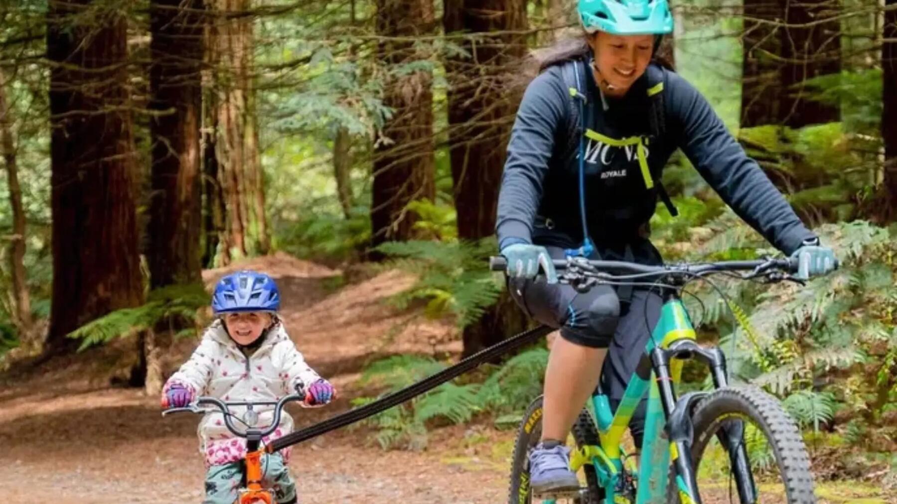 Mama z córeczką na rowerach połączonych za pomocą holu rowerowego jadące po lesie w kaskach rowerowych