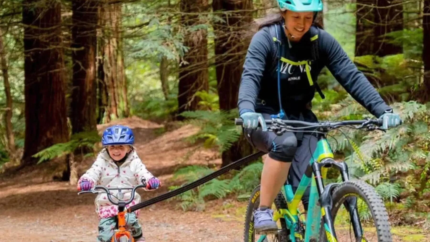 Mama z córeczką na rowerach połączonych za pomocą holu rowerowego jadące po lesie w kaskach rowerowych