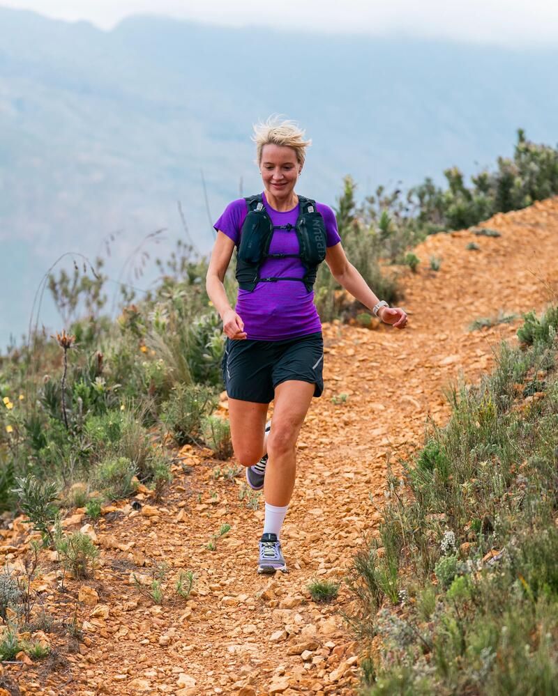 Frau in lila Shirt joggt auf schmalem, steinigen Pfad in grüner, hügeliger Landschaft.