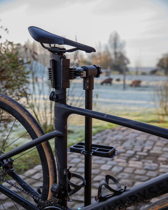 Fahrrad ist an der Sattelstütze in Montageständer eingespannt; im Hintergrund gepflasterter Weg und Kuhweide