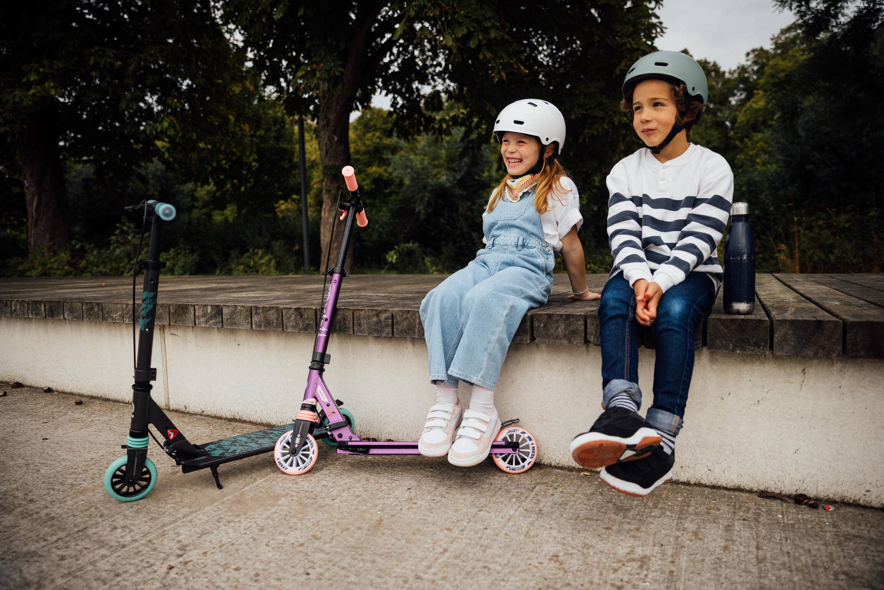 Zwei Kinder mit Fahrradhelmen sitzen gut gelaunt auf einer Holzbank im Park, während vor ihnen zwei bunte Kinder-Scooter parken. Das Bild vermittelt Spaß, Bewegung an der frischen Luft und die Wichtigkeit von Schutzausrüstung beim Rollerfahren.