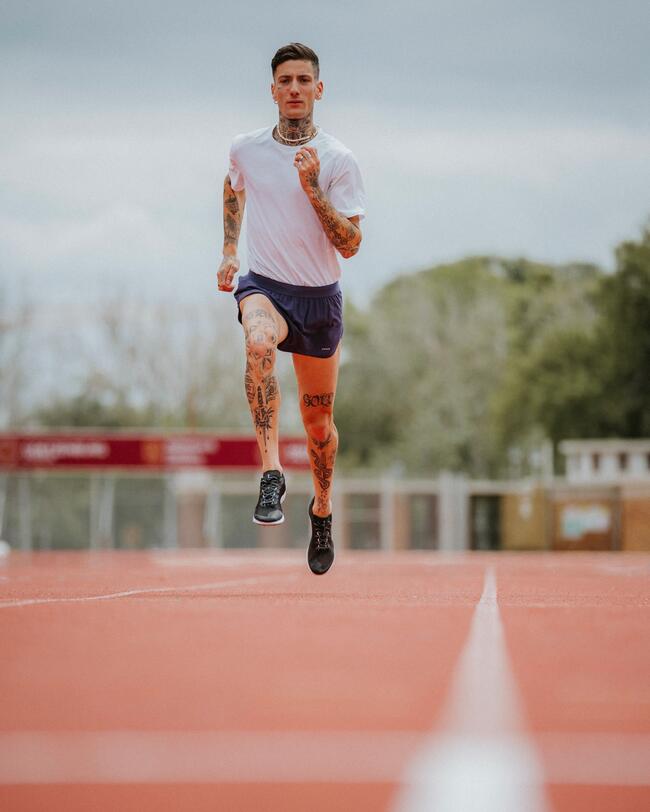Mann mit Tattoos läuft auf roter Laufbahn, trägt weißes Shirt und blaue Shorts, Himmel bewölkt, Bäume im Hintergrund.