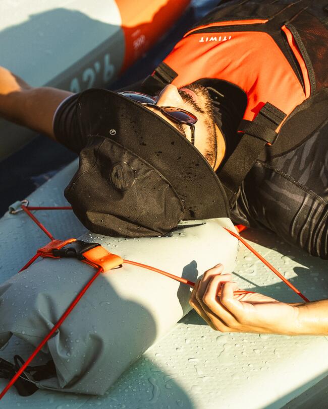 Person mit schwarzem Hut und Sonnenbrille entspannt auf einem Kajak, orange Schwimmweste, Wasserperlen auf grauer Oberfläche.