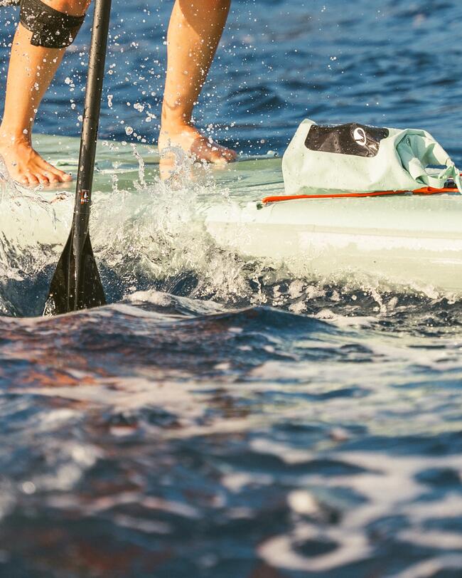 Person steht auf Paddleboard im blauen Wasser, Sonnenlicht reflektiert, Wasser spritzt, Paddel in Bewegung.