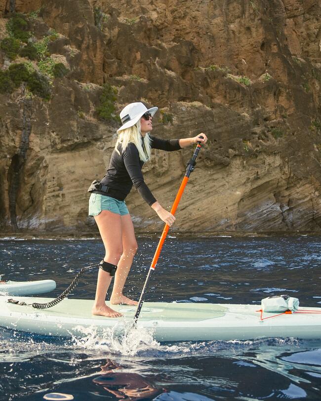 Frau paddelt stehend auf blauem Wasser, trägt Hut, Sonnenbrille und Shorts, Felswand im Hintergrund.
