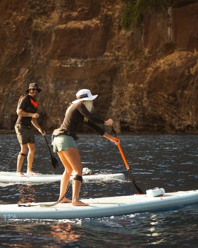 Zwei Personen paddeln auf Stand-Up-Paddleboards vor einer felsigen Küste bei Sonnenschein.