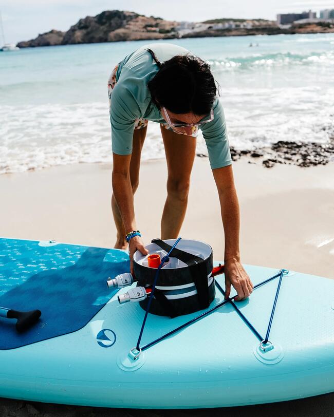 Frau am Strand bereitet ein türkisfarbenes Stand-Up-Paddleboard mit schwarzem Eimer bei sonnigem Wetter vor.