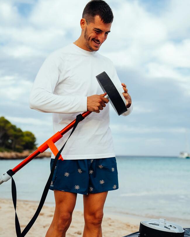 Mann am Strand in blauer Badehose mit Palmenmuster, hält Paddel, Hintergrund Meer und bewölkter Himmel.