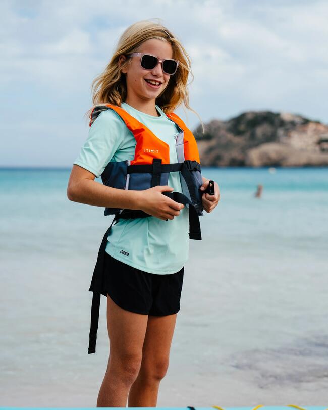 Mädchen mit Sonnenbrille und Rettungsweste am Strand, hellblauer Himmel, türkisfarbenes Wasser, entspannte Atmosphäre.