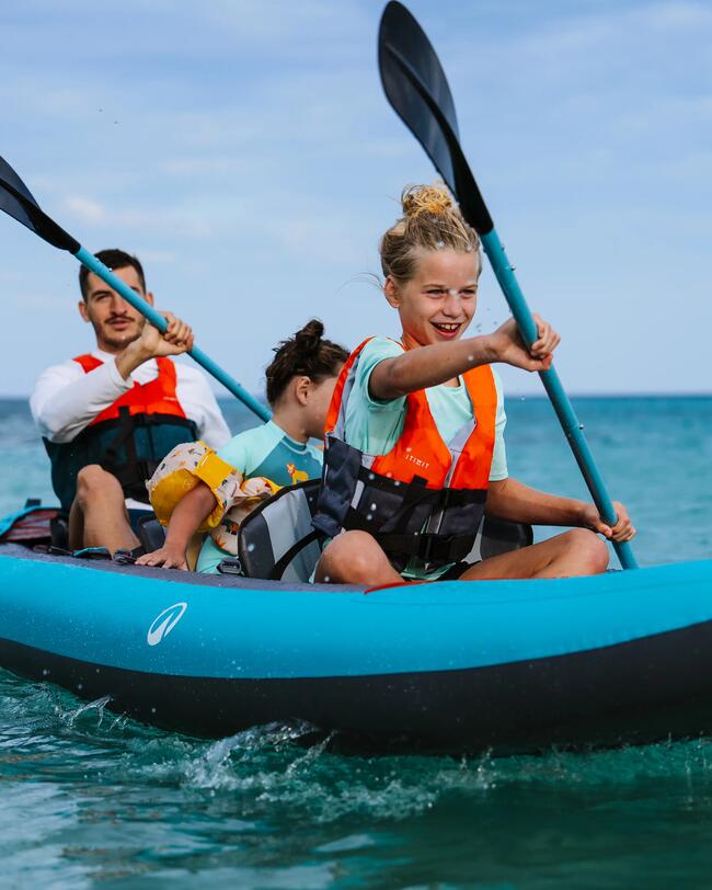 Familie paddelt im blauen Kajak auf ruhigem Meer, leuchtend orangefarbene Schwimmwesten, klarer Himmel.
