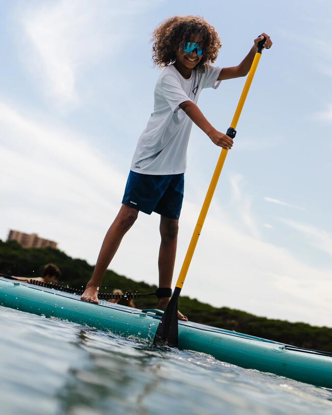 Kind auf türkisfarbenem Paddleboard, blauer Himmel, weiße Wolken, gelbes Paddel, lockiges Haar, entspannte Sommeratmosphäre.