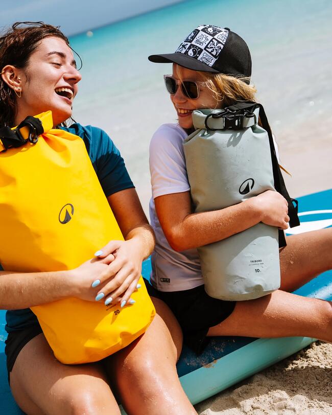 Zwei lachende Personen am Strand, halten gelbe und graue wasserdichte Taschen, tragen Sonnenbrillen und Mützen.