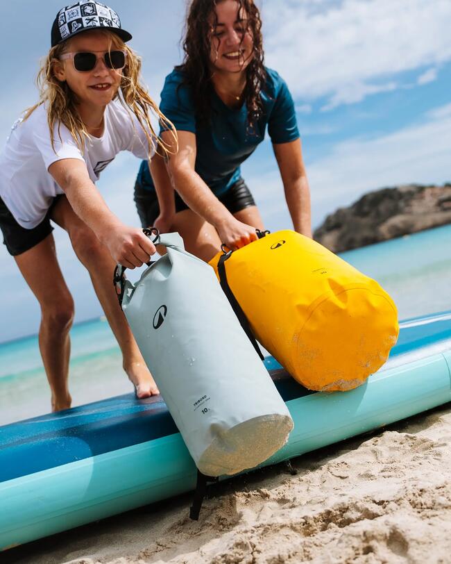 Zwei lächelnde Personen halten gelben und grauen Sack am Strand, blaues Surfbrett im Hintergrund, sonniger Tag.