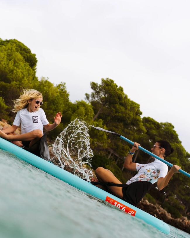Zwei Personen auf einem blauen Paddelboard, lachend im spritzenden Wasser, umgeben von grünen Bäumen und grauem Himmel.