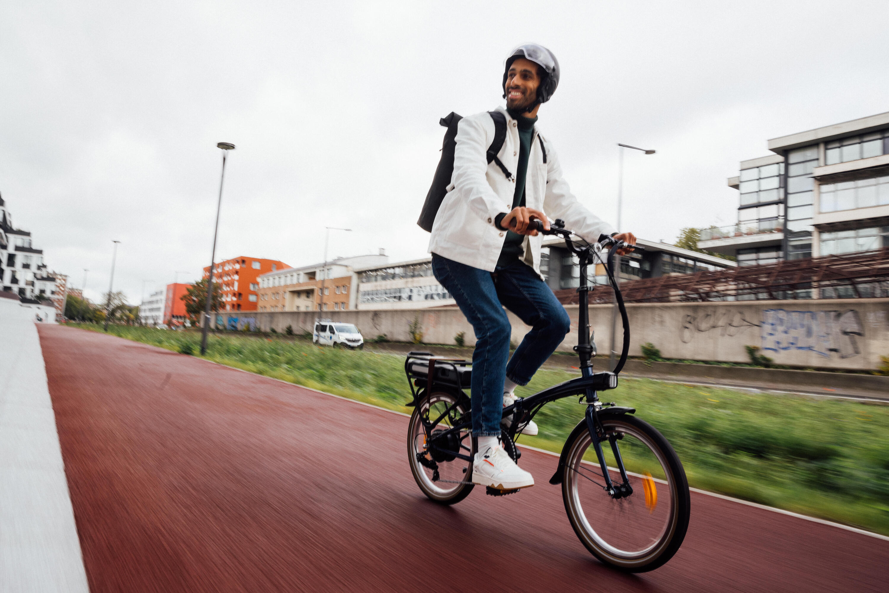 Ein Mann fährt mit einem Helm und Rucksack lächelnd auf einem schwarzen E-Klapprad einen rothen Radweg entlang. Die Szene strahlt dynamische, moderne und flexible Mobilität im städtischen Raum aus.