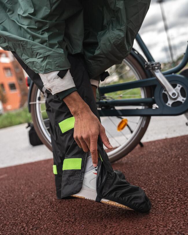Hand zieht schwarzen Regenüberschuh über weißen Sneaker, grüner Mantel und Fahrrad im Hintergrund, braune Bodenfläche.