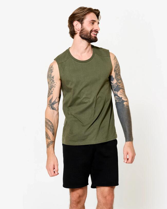 Bärtiger Mann mit tätowierten Armen in einem olivgrünen, ärmellosen Shirt und schwarzen Shorts.