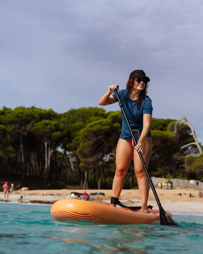 Person beim Stand-Up-Paddling auf türkisfarbenem Wasser, trägt Hut, im Hintergrund Bäume und Sandstrand, bewölkter Himmel.