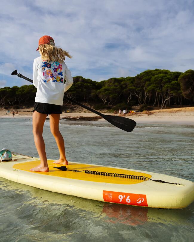 Person paddelt auf gelbem SUP-Board im klaren Wasser, trägt orangenen Hut, bewaldeter Strand im Hintergrund, blauer Himmel.