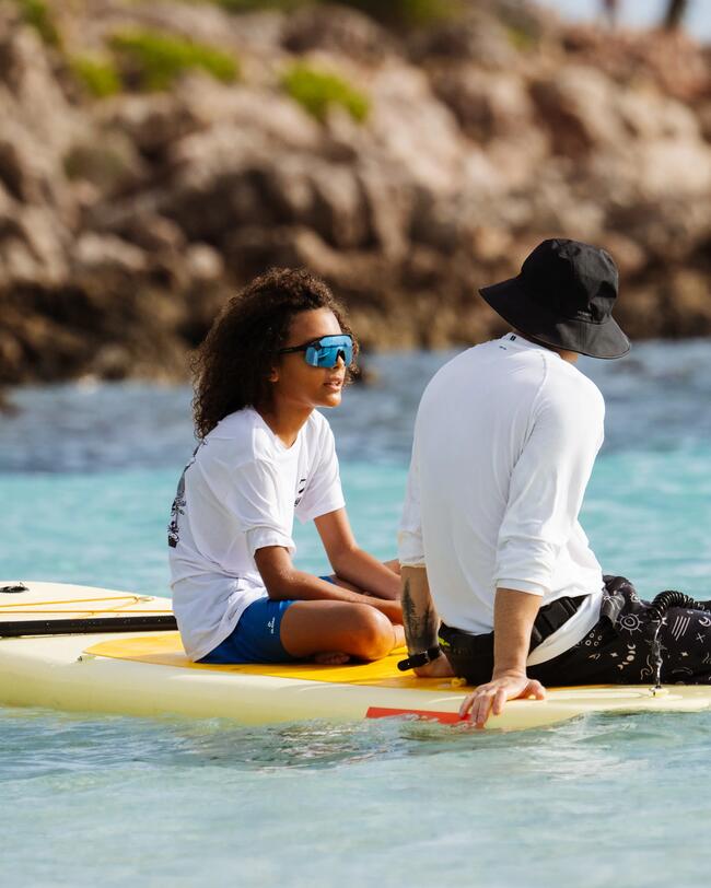 Zwei Personen auf gelbem Paddleboard im klaren, blauen Wasser, Felsen im Hintergrund, Person mit Hut, ruhige Szene.