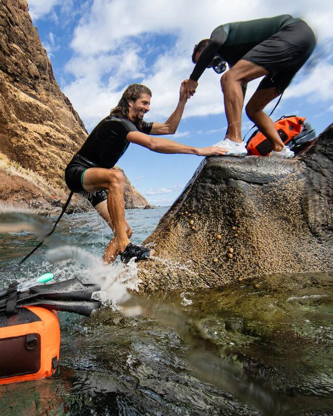 Mann hilft zweitem Mann beim Aussteigen aus dem Wasser auf einen Felsen, Neoprenshorty, Aquaschuhe Sneaker