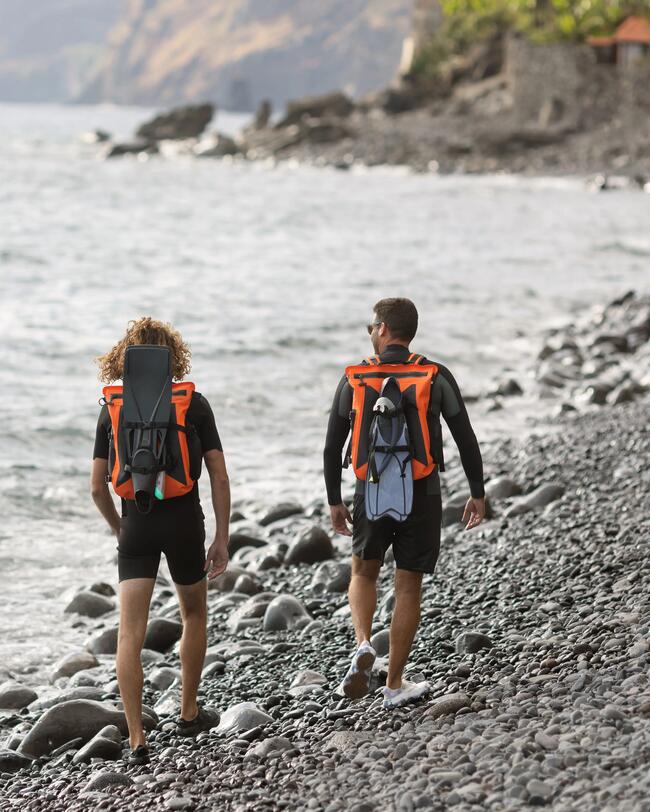 Zwei Personen mit orangefarbenen Rucksäcken wandern über schwarzen Kiesstrand, Meer und Felsen im Hintergrund.