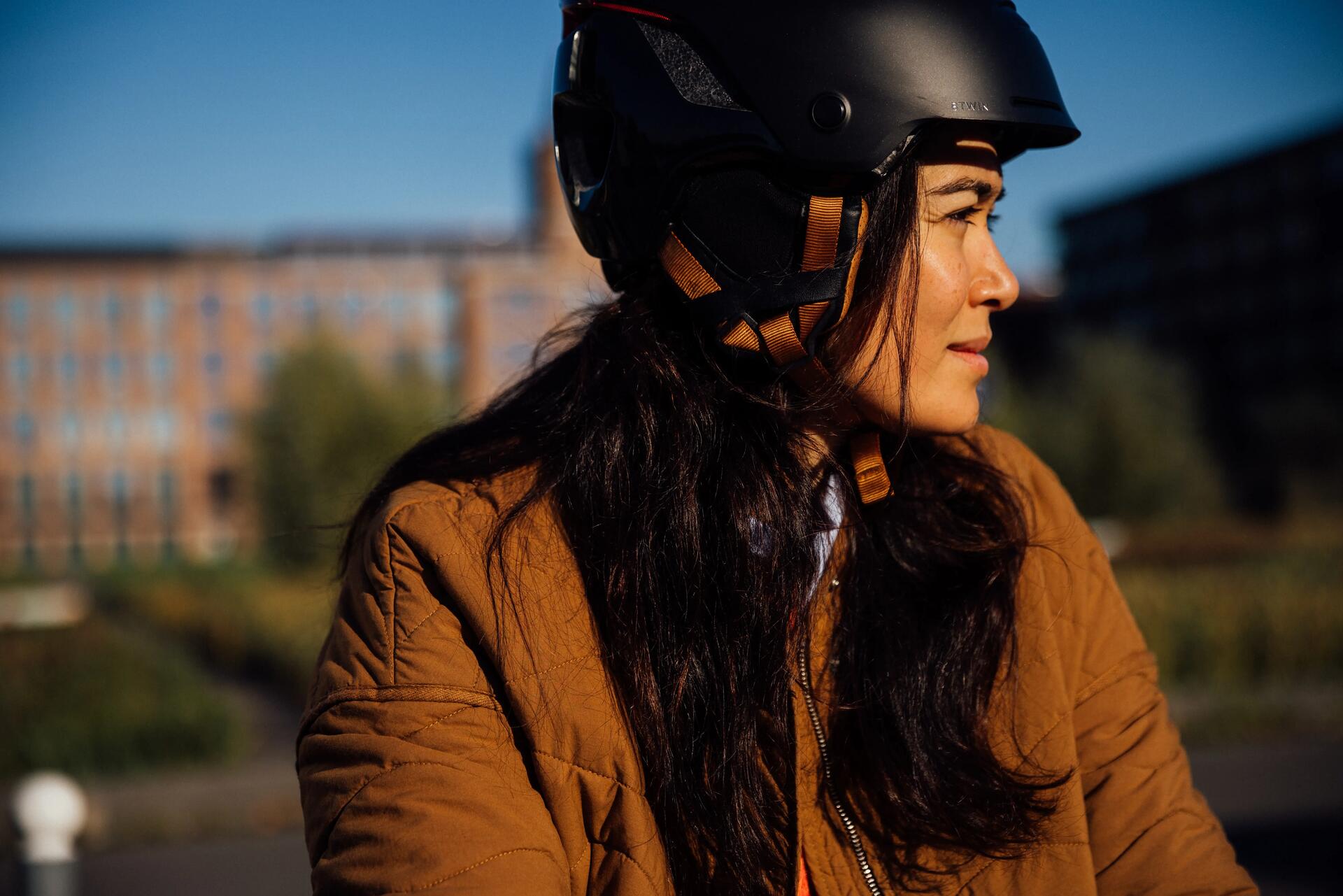 Frau mit brauner Jacke und schwarzem Helm vor unscharfem Gebäude, warmes Sonnenlicht.