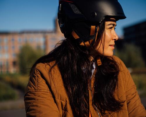 Frau mit brauner Jacke und schwarzem Helm vor unscharfem Gebäude, warmes Sonnenlicht.
