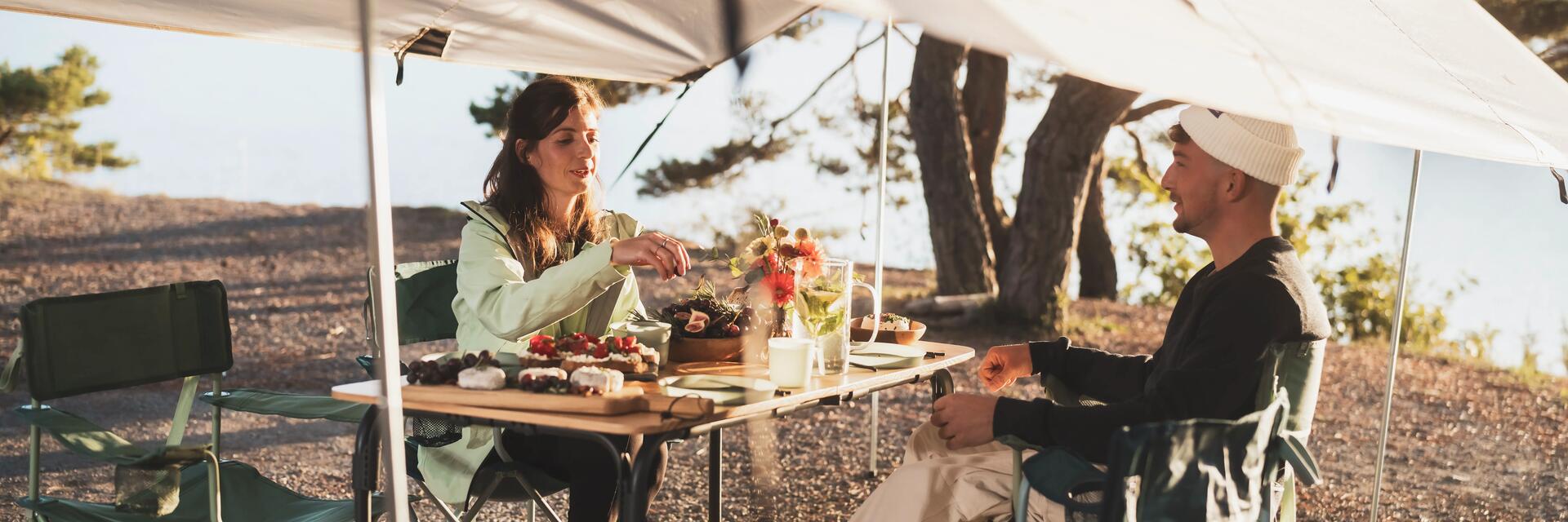 Zwei Personen sitzen gemeinsam am Campingtisch unter einem Pavillon beim entspannten Frühstück-