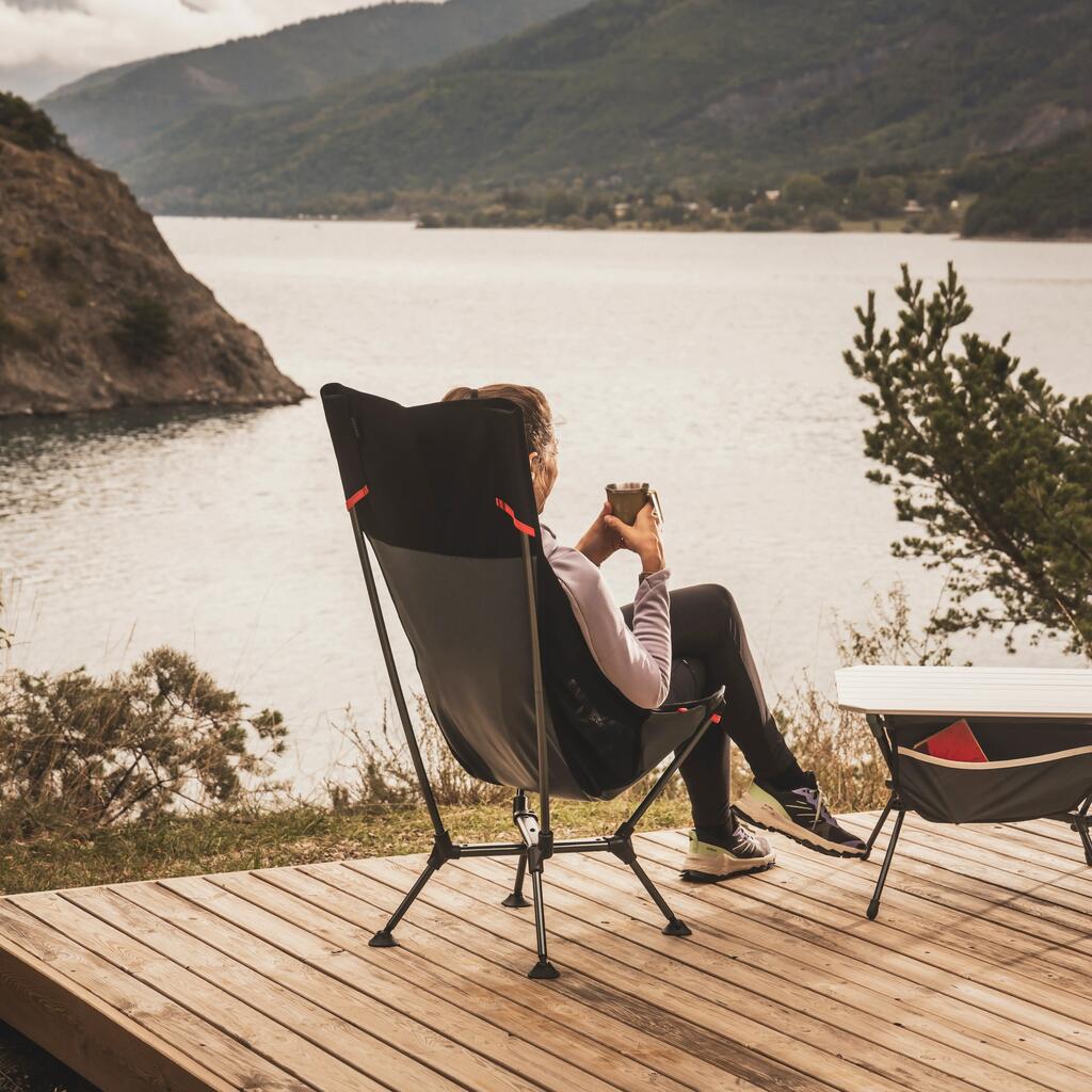 Osoba sedi u tamnozelenoj Quechua sklopivoj stolici na drvenoj terasi i uživa u pogledu na mirno jezero i planine.