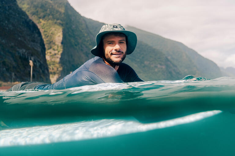 Surfer wearing a green surf hat floating on a surfboard in the ocean.