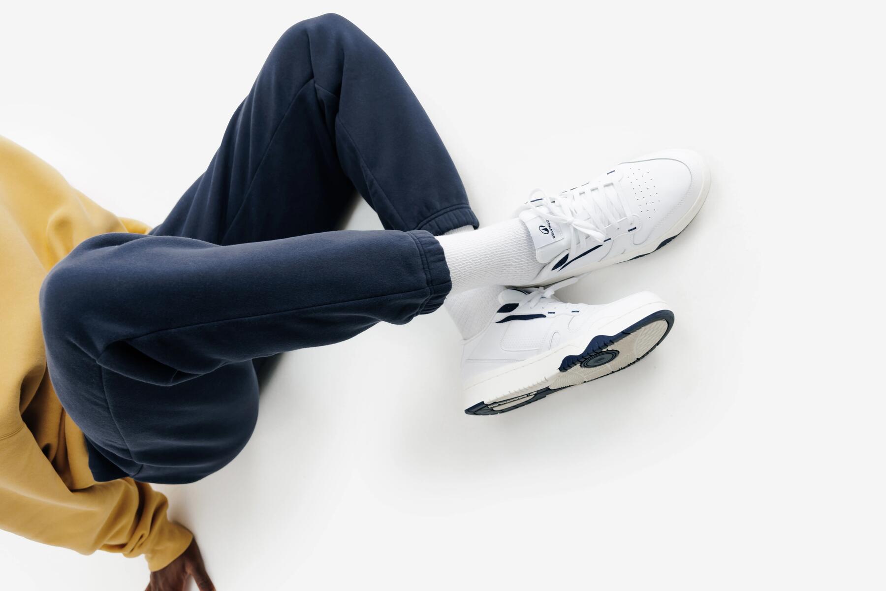 Mężczyzna siedzący ze skrzyżowanymi nogami w granatowych spodniach dresowych i białych sneakersach