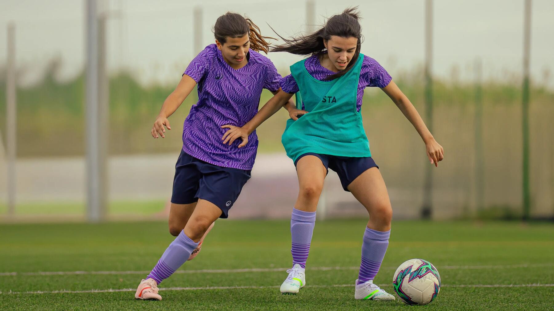 Zwei junge Frauen spielen Fußball auf einem Kunstrasenplatz im Freien, während sie im Zweikampf um den Ball kämpfen.
