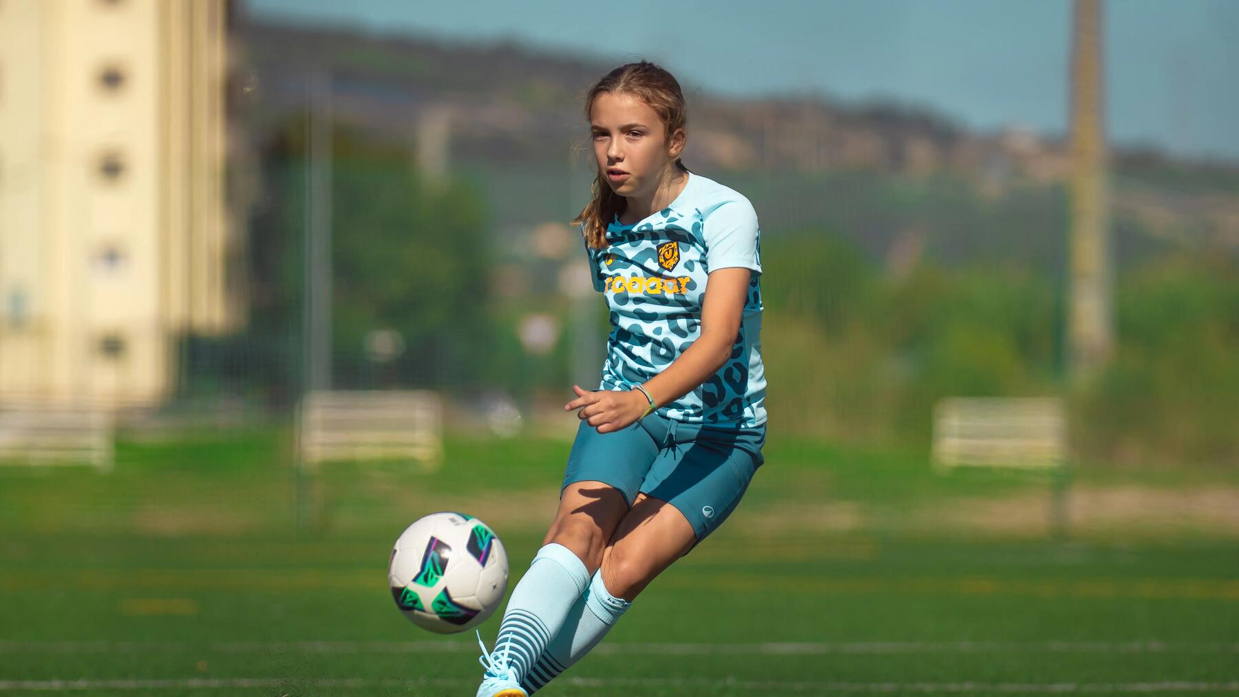 Mädchen im türkisen Outfit schießt einen Fußball auf Fußballplatz. Berglandschaft im Hintergrund.
