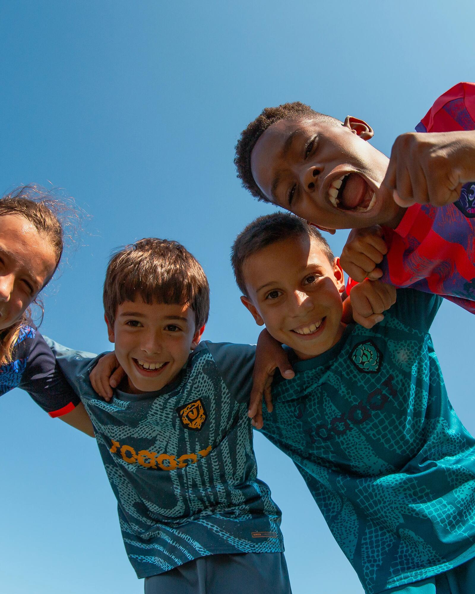 Vier jubelnde Kinder in Sporttrikots blicken aus einer Froschperspektive lächelnd in die Kamera vor blauem Himmel.