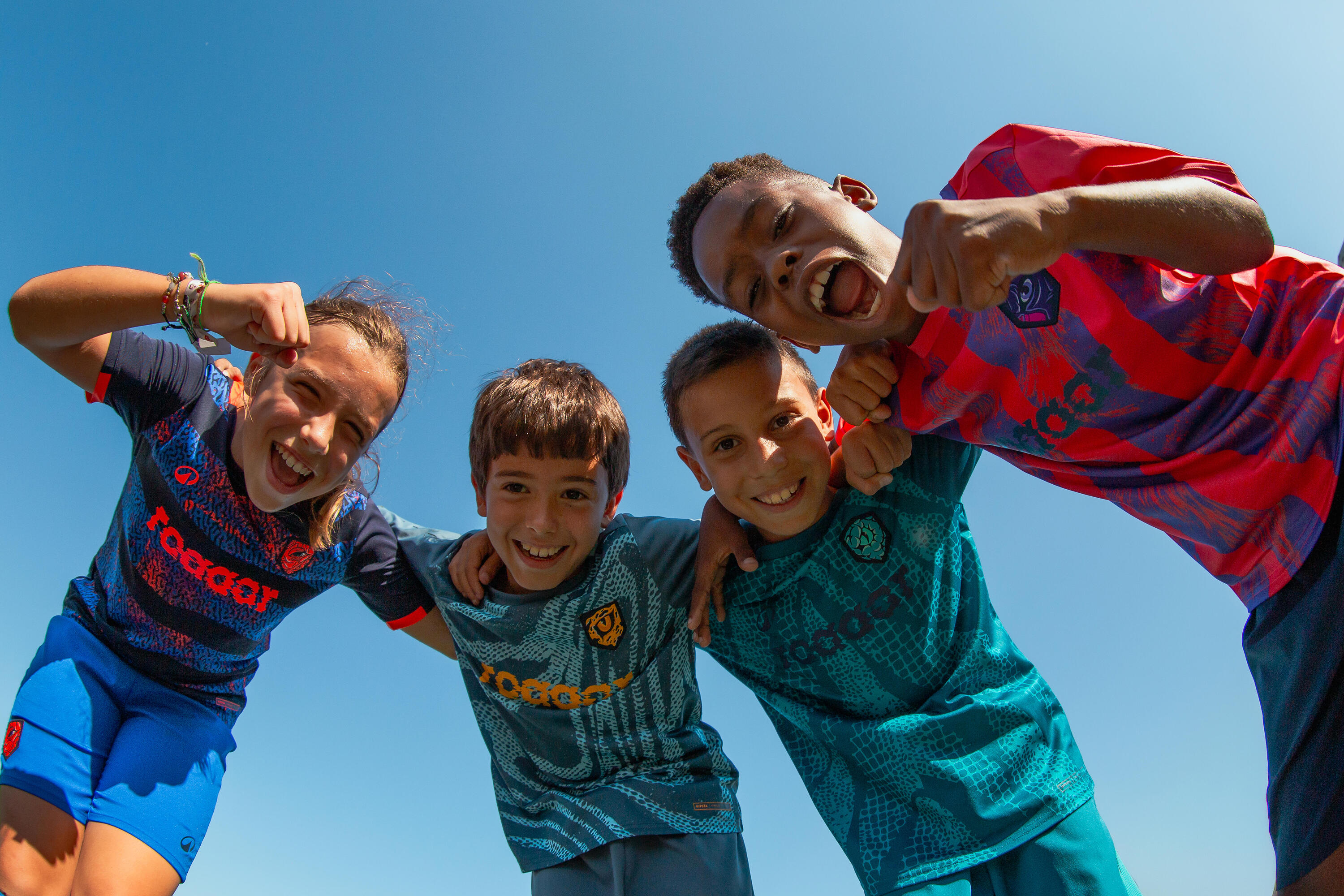 Kinder brüllen lachend in Fußballtrikots in die Kamera. Blauer Himmel.