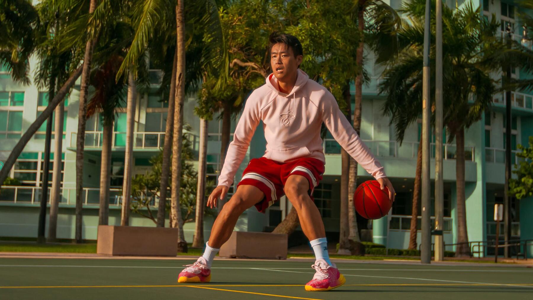 Person spielt Basketball auf einem Platz, trägt roten Shorts und Hoodie; umgeben von Palmen und modernen Gebäuden.