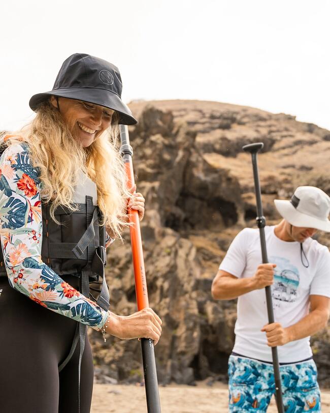 Frau und Mann mit Paddeln am Strand, lächeln, tragen Sonnenhüte und bunte Outfits, Felsen im Hintergrund.
