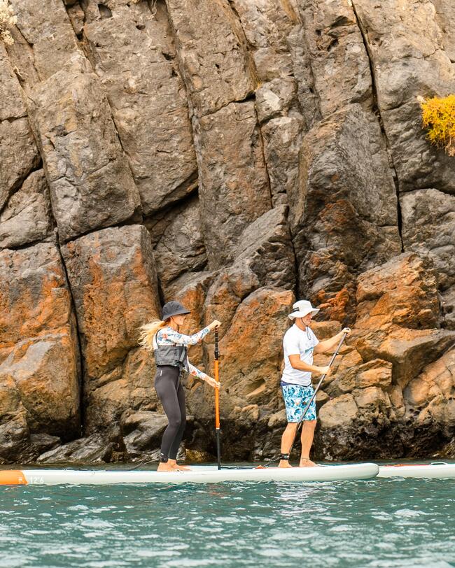 Zwei Personen auf Paddleboards vor grauen, felsigen Klippen, türkisfarbenes Wasser, sportliche Kleidung, helle und dunkle Farben.