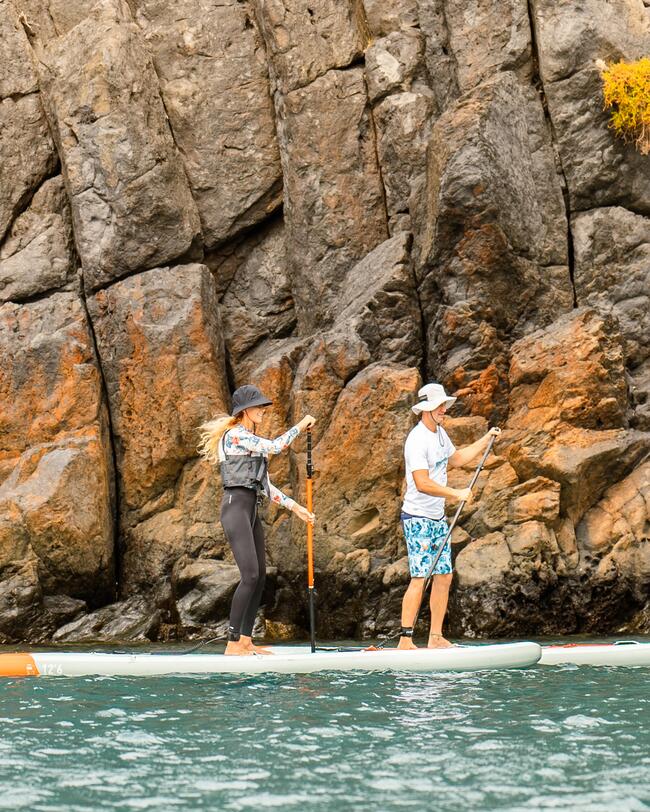 Zwei Personen auf Paddleboards vor einer felsigen, braunen und grauen Küste mit spritzendem blauen Wasser.